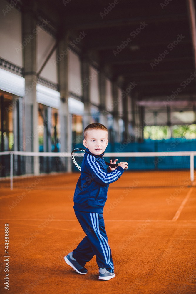 Obraz premium little boy on the yellow, sandy tennis court stands with racket in blue tracksuit