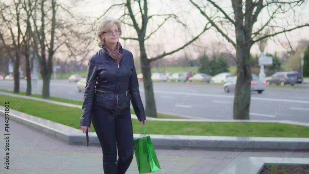 An attractive middle-aged woman is walking down the street. She is holding a mobile phone and a package. Slow-motion An attractive middle-aged woman is walking down the street. She is holding a mobile phone and a package. Slow-motion