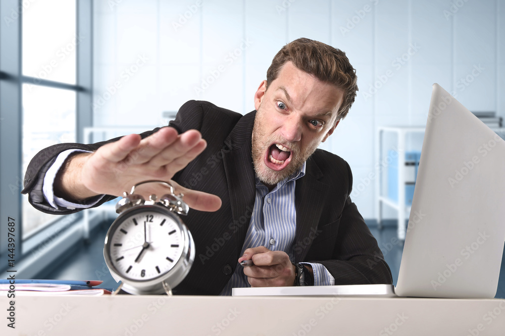 angry exploited businessman at office desk stressed and frustrated with computer laptop and alarm clock angry exploited businessman at office desk stressed and frustrated with computer laptop and alarm clock