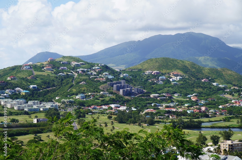 A view over St. Kitts Island with residential area and road on the foreground and lush green hills on the background.