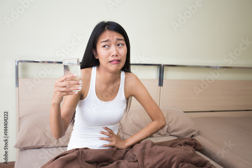 Beautiful cute young woman touches her temple and feels a strong stomachache holding a glass of water in her hand photo