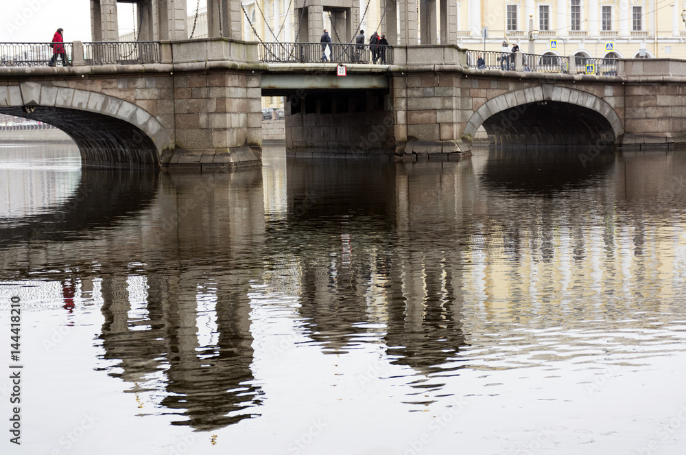 Lomonosov bridge reflected in the water of the Fontanka river, on the bridge are people, cloudy day