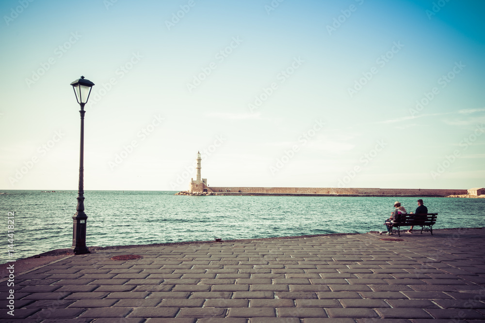 Couple relaxes on a bench at the old harbor of Chania, retro style, Crete, Greece.