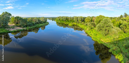 Panorama of river valley with trees and bush growth on riverside reflecting in calm water. Berezichi, Kaluzhskaya region, Russia. 