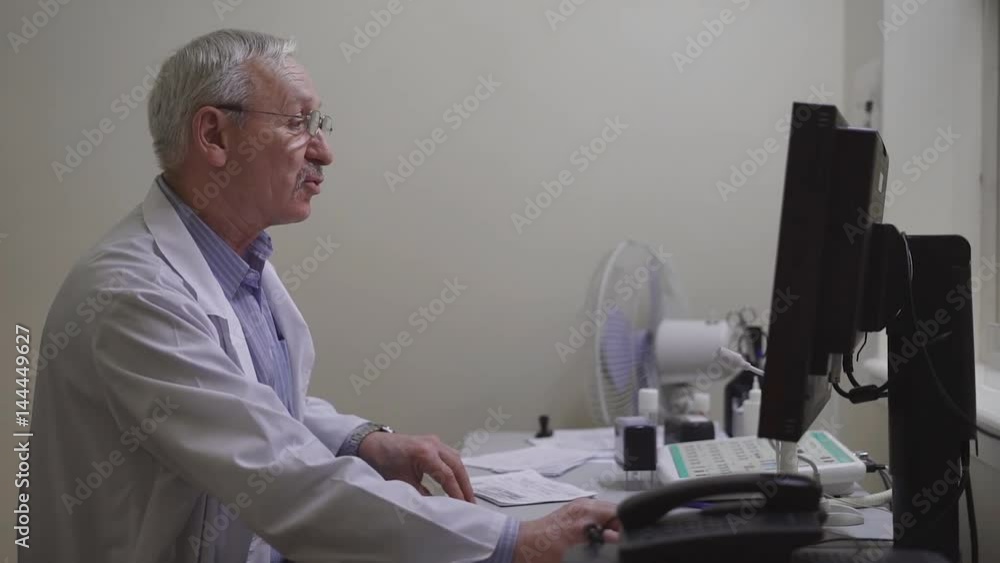 An adult doctor in a dressing gown makes an anamnesis of a patient of a private clinic to make the results in the medical record of a single computer base of a polyclinic An adult doctor in a dressing gown makes an anamnesis of a patient of a private clinic to make the results in the medical record of a single computer base of a polyclinic