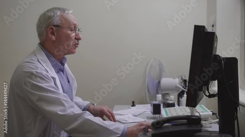 An adult doctor in a dressing gown makes an anamnesis of a patient of a private clinic to make the results in the medical record of a single computer base of a polyclinic photo