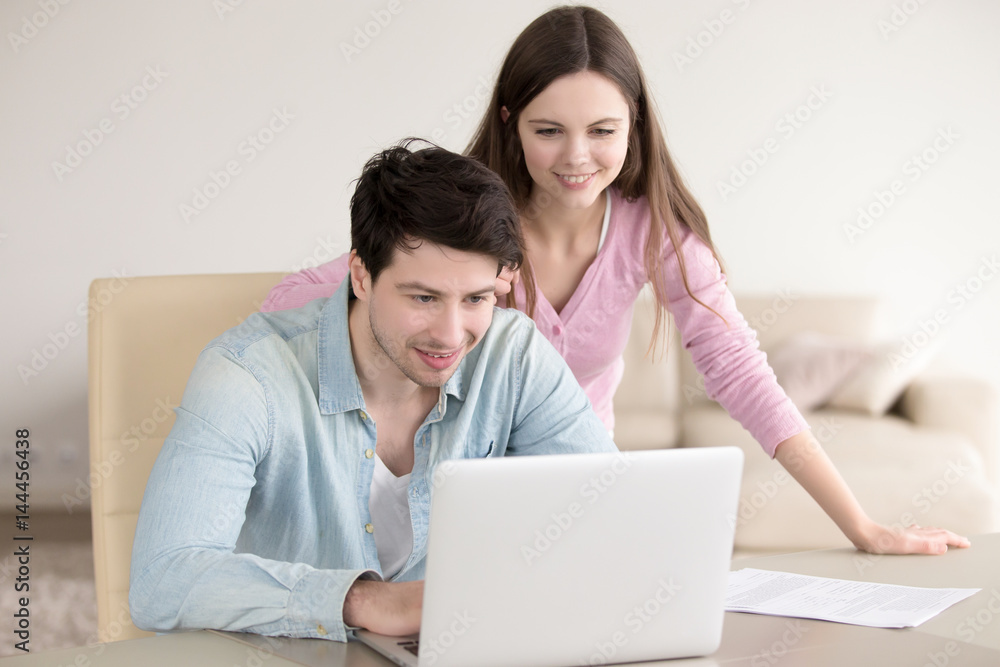 Cheerful young couple using laptop, man and woman discussing mortgage, investment planning, calculating and paying bills, analyzing their finances with documents to save money, working with computer