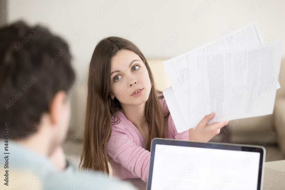 Puzzled young lady holding documents and showing papers to male colleague, asking to choose the best offer. Young couple discussing bills, planning family budget, having problem with documentation Puzzled young lady holding documents and showing papers to male colleague, asking to choose the best offer. Young couple discussing bills, planning family budget, having problem with documentation
