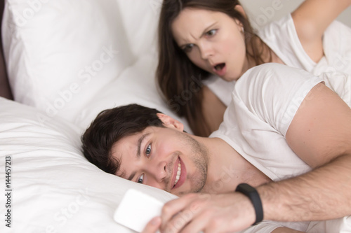 Surprised woman looking at her boyfriends mobile phone, when he texting or reading messages lying with his girlfriend in bed. Wife caught husband watching pornographic videos on smartphone