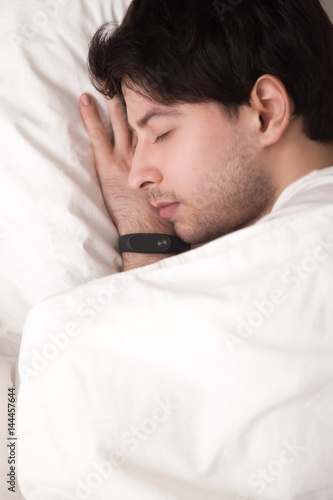 Close up vertical top view of man sleeping on white comfortable bed, wearing digital smartwatch or wristband tracker for monitoring and controlling his sleep. Keeping fit and healthy, time management