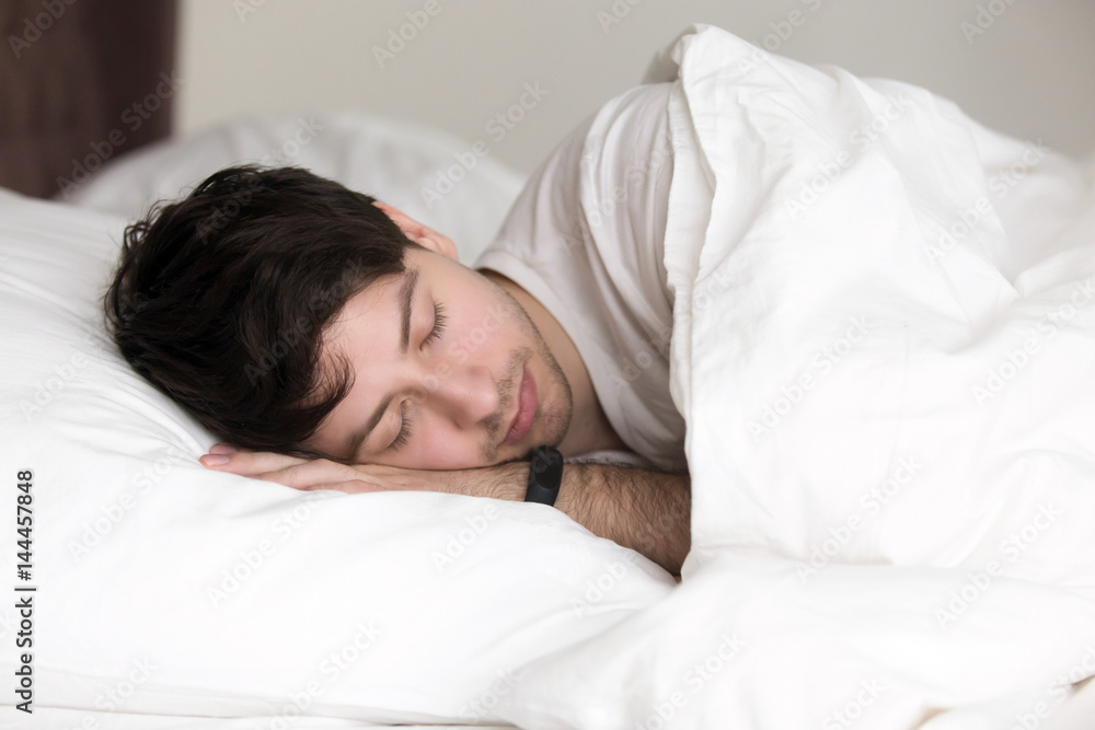 Young handsome guy sleeping in soft bed under blanket, man asleep wearing activity tracker with sleep monitoring features on his wrist to feel full of energy after waking up Young handsome guy sleeping in soft bed under blanket, man asleep wearing activity tracker with sleep monitoring features on his wrist to feel full of energy after waking up