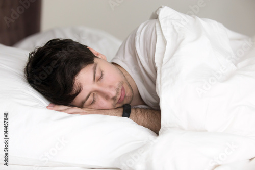 Young handsome guy sleeping in soft bed under blanket, man asleep wearing activity tracker with sleep monitoring features on his wrist to feel full of energy after waking up