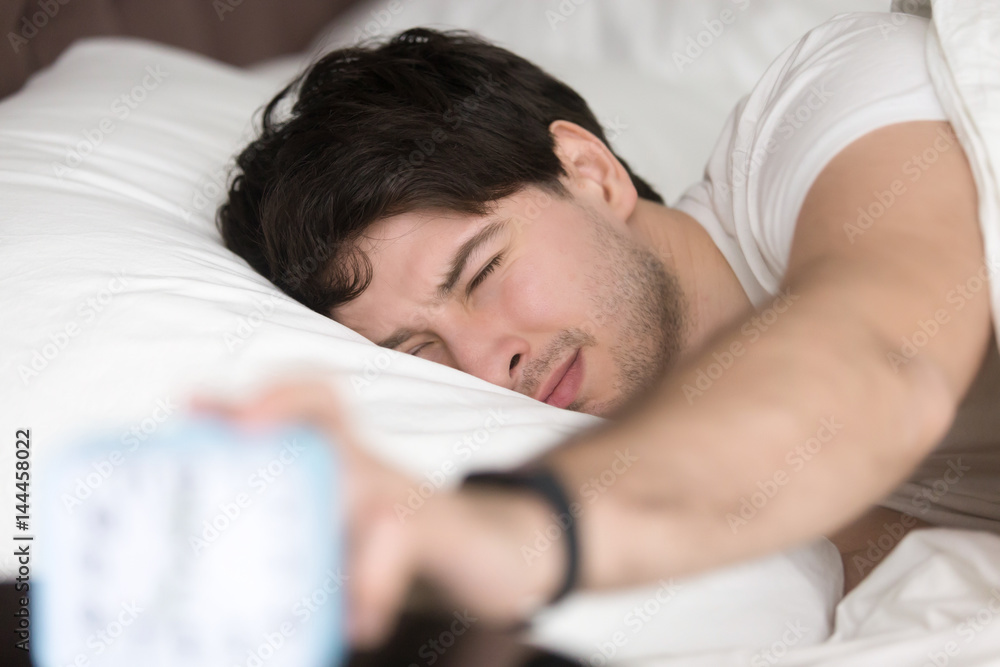 Sleepy guy waking up early after hearing alarm clock signal on monday morning, with eyes still closed reaching button on the clock to turn it off, feeling tired and does not want to get up Sleepy guy waking up early after hearing alarm clock signal on monday morning, with eyes still closed reaching button on the clock to turn it off, feeling tired and does not want to get up