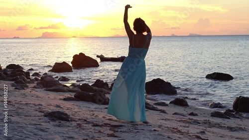 Happy caucasian woman in a long dress walks on the beach on a colourful sunset background
