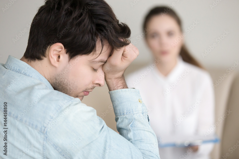 Young worried guy touching his forehead, having a headache or migraine, overworked businessman feeling tired, stressed and exhausted, female doctor, nurse or colleague standing in the background Young worried guy touching his forehead, having a headache or migraine, overworked businessman feeling tired, stressed and exhausted, female doctor, nurse or colleague standing in the background