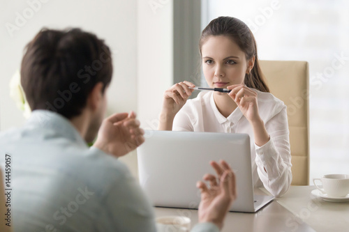 Young businesswoman looking serious and thoughtful, holding a pen near her mouth, listening attentively to self introduction of a male candidate during job interview or considering business offer photo