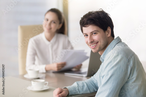 Smiling guy sitting at office desk opposite female human resources manager, looking at camera, apply for a job at successful big company, young entrepreneur promoting his services, health insurance