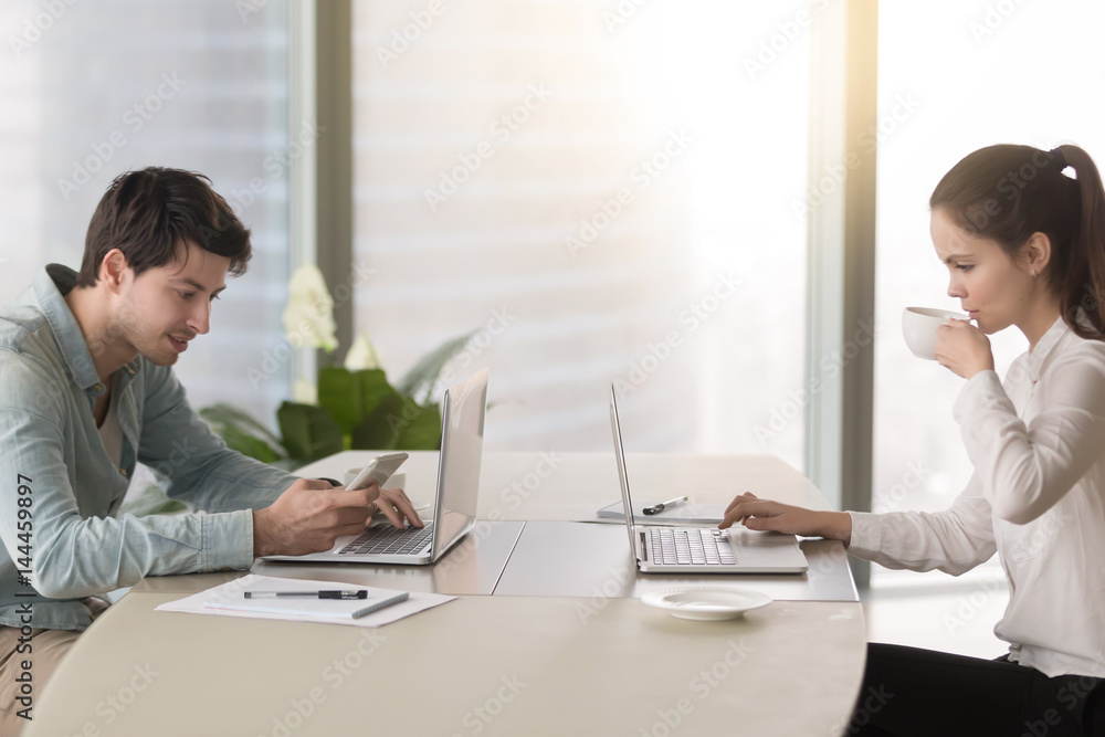Side view of young woman working with laptop enjoying her coffee, man sitting opposite using mobile phone and notebook computer, inefficient workers, coffee break or lunch time at the workplace Side view of young woman working with laptop enjoying her coffee, man sitting opposite using mobile phone and notebook computer, inefficient workers, coffee break or lunch time at the workplace