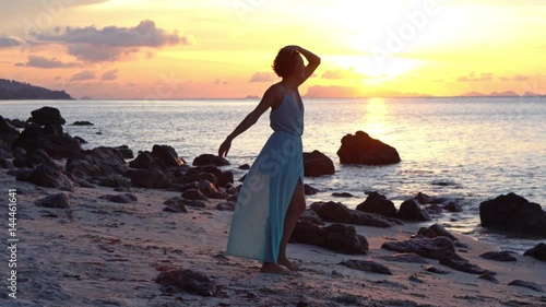 Happy caucasian woman in a long dress walks on the beach on a colourful sunset background