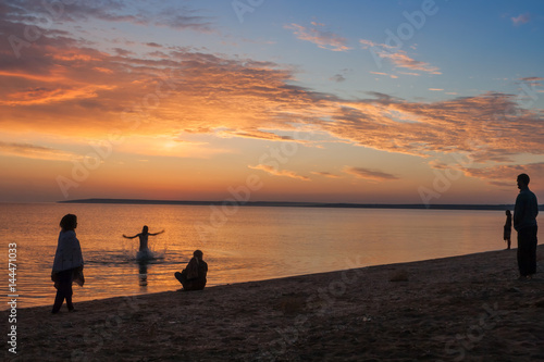Beautiful colorful summer sea sunrise landscape with amazing colorful clouds in a blue sky and unrecognizable people silhouettes waiting for the Sun. One man is splashing and jumping in water. In