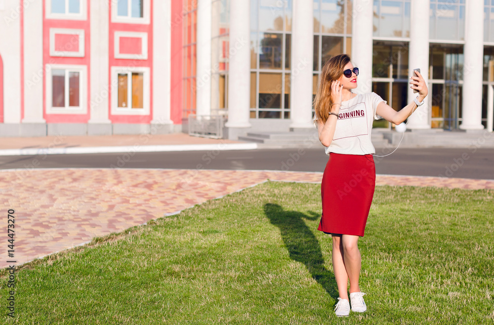 Full-height portrait of a standing smiling young girl holding a smartphone and making a selfie. Girl wears a white t-shirt, red skirt and dark sunglasses. Girl is in the city center.