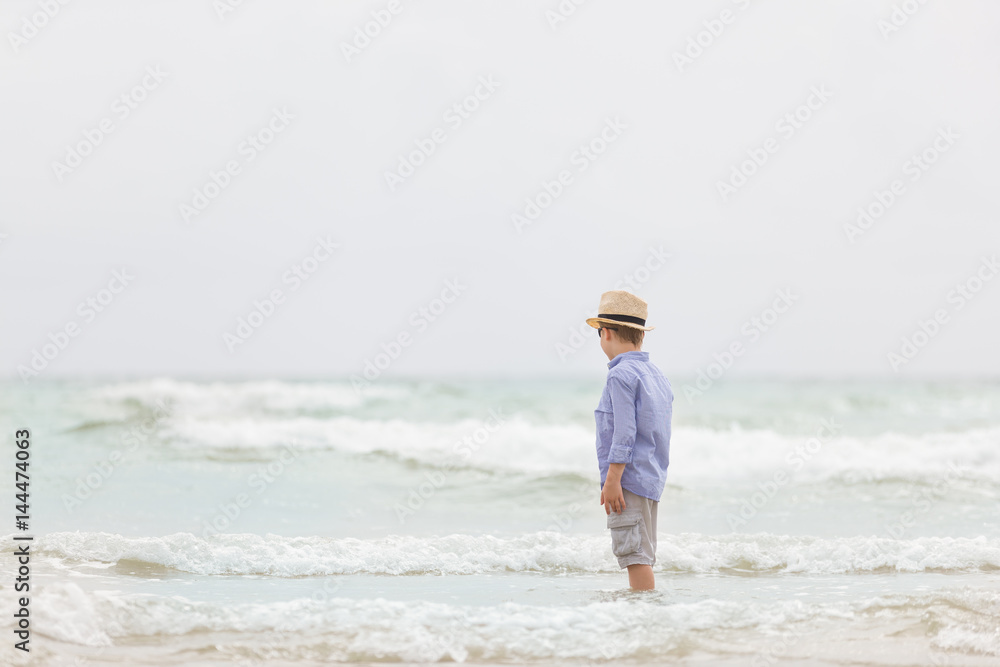 Adorable kid boy in straw hat and sun glasses walking on ocean beach and playing with waves. Vacations by the sea. Outdoor.