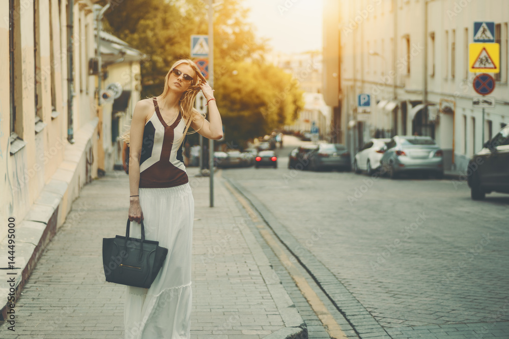 Attractive adult tall caucasian woman in long white skirt, sunglasses and with bag is fixing her hair while standing on street during sunny summer day with copy space for your advertising message