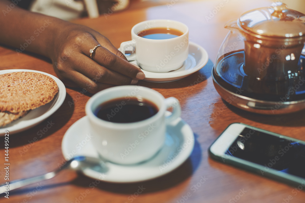 Fototapeta Hand of young black girl touching cup with delicious tea on table with sesame biscuits, teapot, one more cup and smartphone, biracial teenager arm holding white saucer while meeting during breakfast