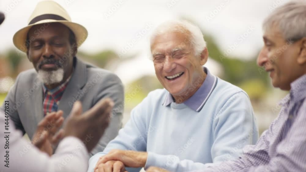  Happy senior male friends chatting & laughing together outdoors in the park