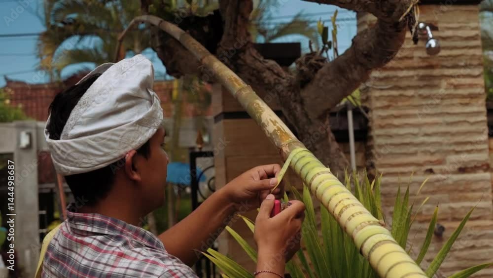 Balinese man making penjor on Galungan Day (Bali holiday ceremony) - covering a bamboo pole with a palm branch ribbon using stapler on sunny day. Close up shot with Sony a7s on slider