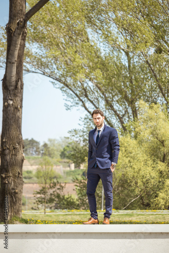 one young man, formal clothes suit tie, posing, looking at camera, standing on wall, nature trees, grass © HD92
