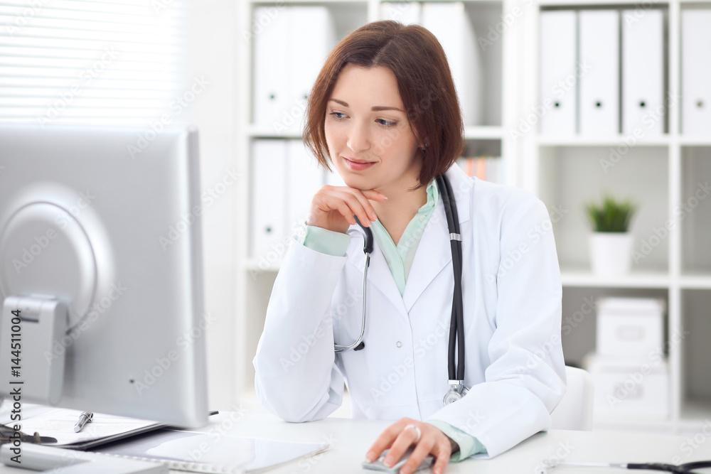 Young brunette female doctor sitting at a desk and working on the computer at the hospital office. Health care, insurance and help concept. Physician ready to examine patient Young brunette female doctor sitting at a desk and working on the computer at the hospital office. Health care, insurance and help concept. Physician ready to examine patient