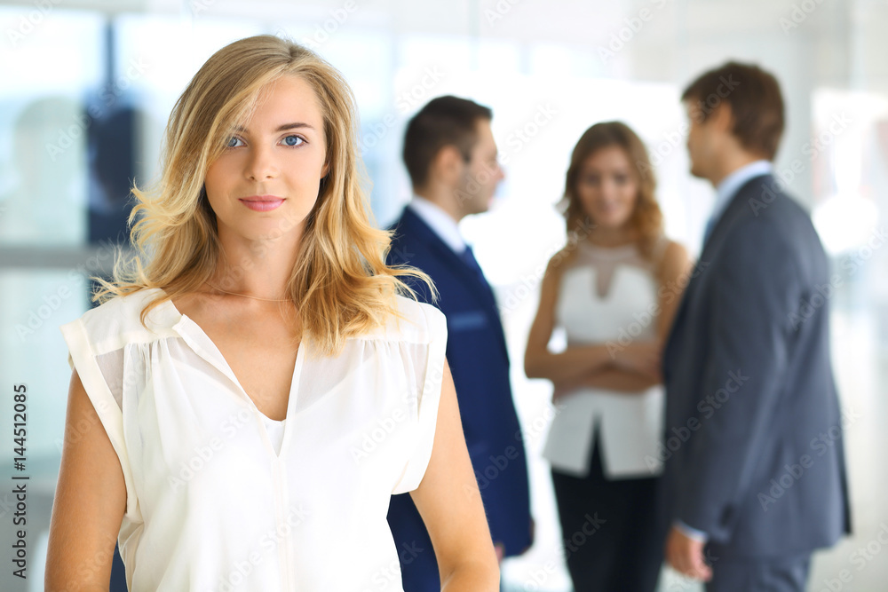 Blonde woman with touchpad computer looking at camera and smiling while business people are shaking hands over office background Blonde woman with touchpad computer looking at camera and smiling while business people are shaking hands over office background