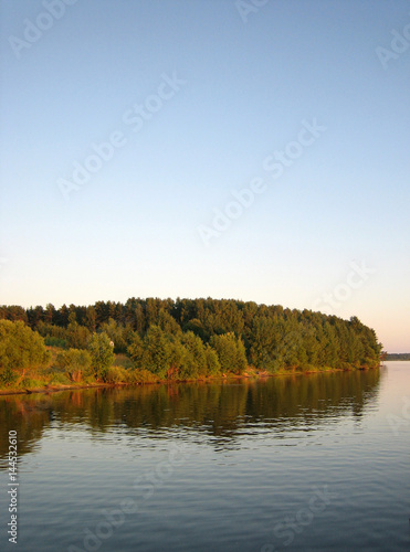 Green autumnal forest near river on blue sky background, vertical view.