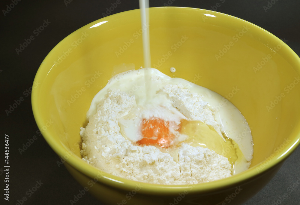 Pouring milk into yellow ceramic mixing bowl with dry flour and egg for making pancakes Pouring milk into yellow ceramic mixing bowl with dry flour and egg for making pancakes