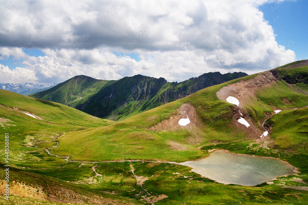 A small lake in the area of alpine meadows in the Caucasus. White clouds in the blue sky.
