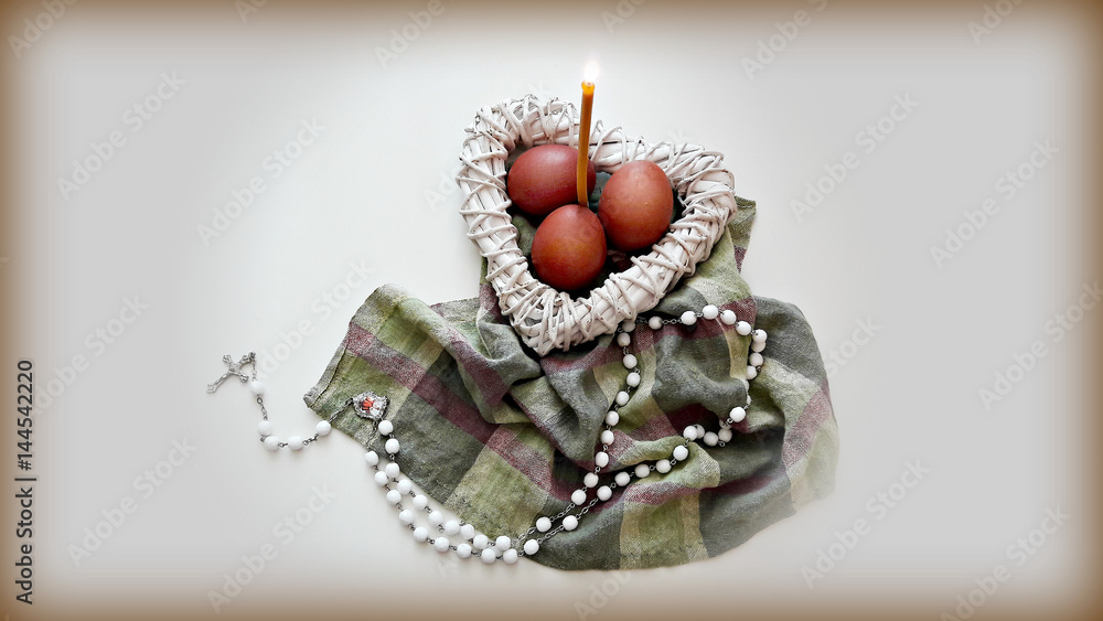 Naklejka premium Easter composition on the table. Colored eggs in a white wicker heart with a burning candle and a cross.