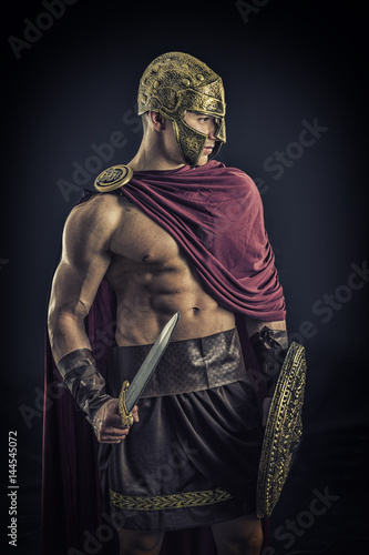 Young handsome muscular man posing in roman or spartan gladiator costume with shield and sword, on black background in studio