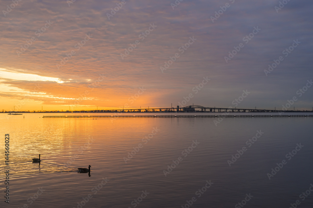 Vibrant sunrise sky and cloudscape reflected in calm lake waters, birds and bridge in silhouette