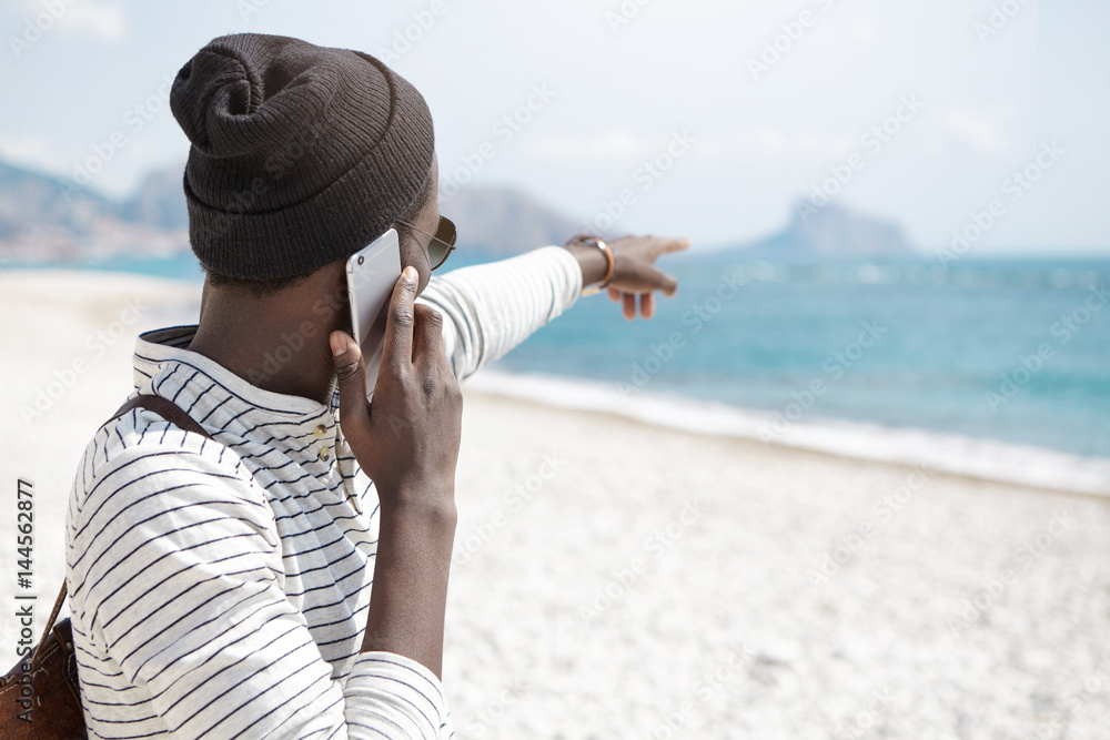 Unrecognizable dark-skinned young male tourist dressed in trendy clothing standing on white sandy beach and pointing his index finger into distance towards ocean while having phone conversation