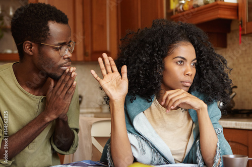 People, relationships problems and divorce. Repentant worried dark-skinned male keeping hands pressed together, begging offended wife to forgive his infidelity, mad woman not looking at him at all photo