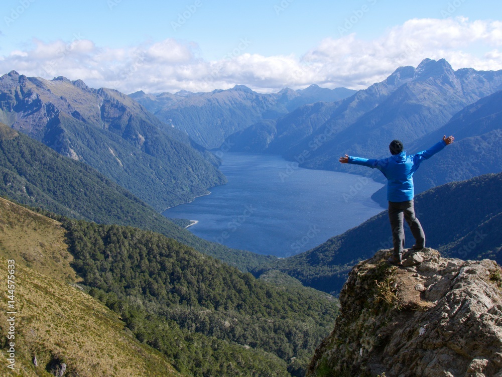 Man standing on mountain ridge with lifted arms looking over a lake