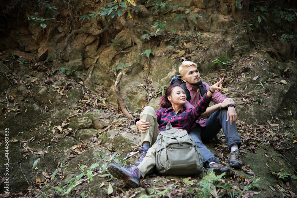 Young tourist couple take a rest near waterfall while hiking in forest looking away in search of attractions. Travel concept.