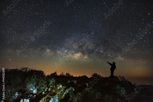 A Man is standing next to the milky way galaxy pointing on a bright star at Doi Luang Chiang Dao with Thai Language top point signs. Long exposure photograph.With grain