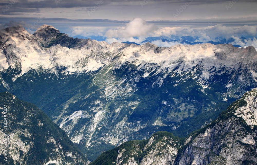Jalovec, Mangart peaks, Mojstrovke ridge, dark green pine forests of Trenta Valley, Julian Alps, Triglav National Park, Slovenia, in the background Gailtal Alps, Carinthia, Austria in clouds