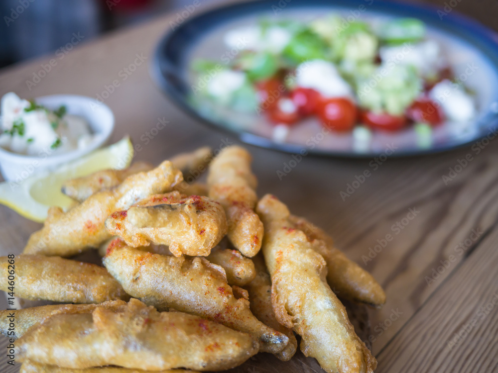 Deep fried whitebait, sprats in batter served with a lemon slice and dip on a wooden table with a tomato, feta cheese and avocado salad. Deep fried whitebait, sprats in batter served with a lemon slice and dip on a wooden table with a tomato, feta cheese and avocado salad.