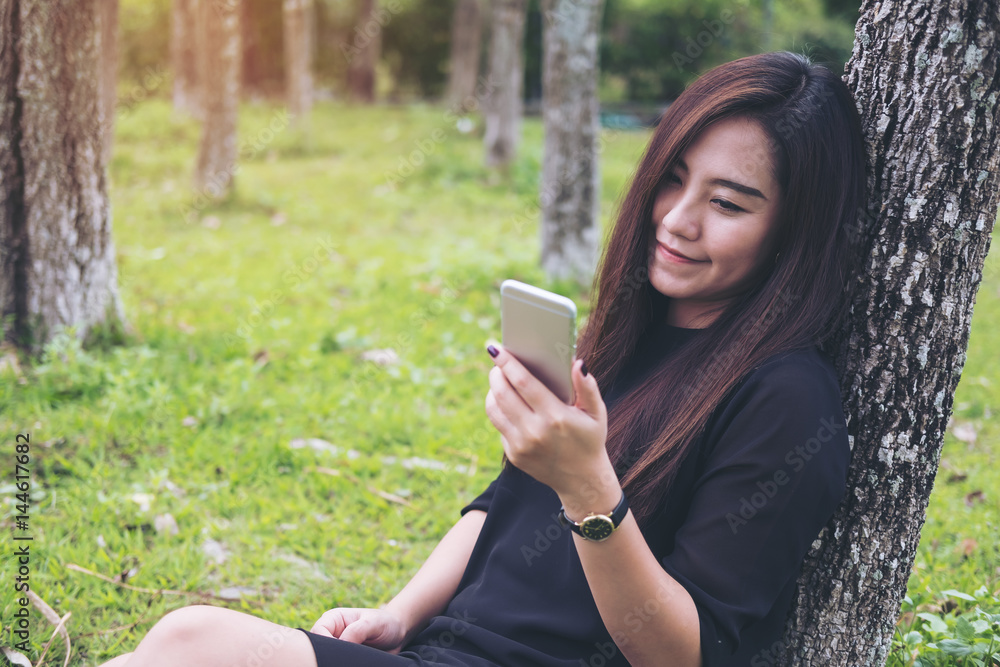 A beautiful Asian woman sitting by the tree and using smart phone with green forest background A beautiful Asian woman sitting by the tree and using smart phone with green forest background