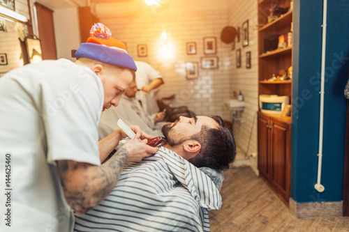 Handsome bearded man, hipster, brunette with beard and moustache has haircut or clippering in hairdressing saloon or barbershop. Barber works with clipper photo