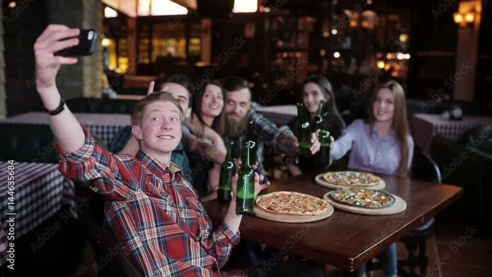 The guy makes a friend company in pizzeria. A company of friends resting in a pizzeria and making selfie on the background of pizza and beer The guy makes a friend company in pizzeria. A company of friends resting in a pizzeria and making selfie on the background of pizza and beer