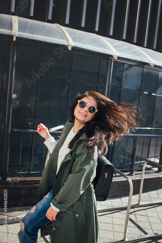 fashion closeup portrait of nice pretty young hipster woman posing in sunglasses Outdoor .Brunette happy girl in green raincoat and pink sneakers walks the street of the city.